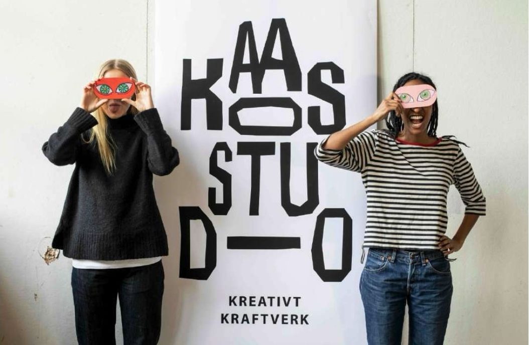 Kaksi henkilöä pitelee värikkäitä silmänaamioita silmiensä edessä KAAOSSTUDIO-julisteen edustalla.